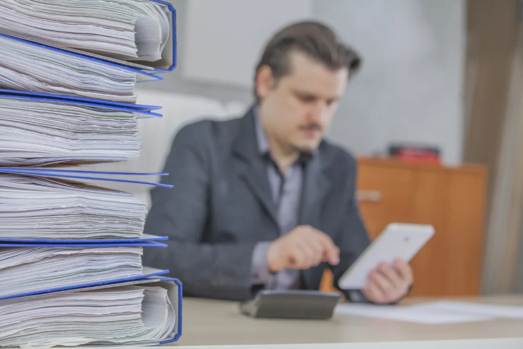 Imagem de um contador fazendo cálculos e a gestão de documentos contábeis diante de uma pilha de pastas.