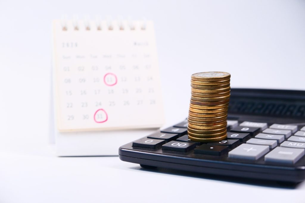 Imagem de uma pilha de moedas sobre uma calculadora e um calendário de fundo, simbolizando o cronograma da Reforma Tributária.
