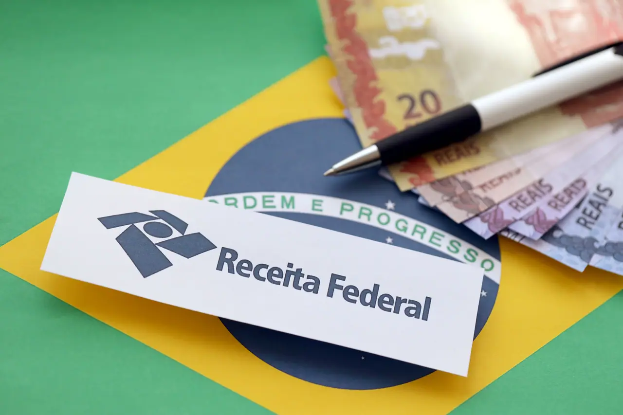 Cédulas de dinheiro e um papel com a logo da Receita Federal sobre a bandeira do Brasil, simbolizando a autorregularização fiscal.