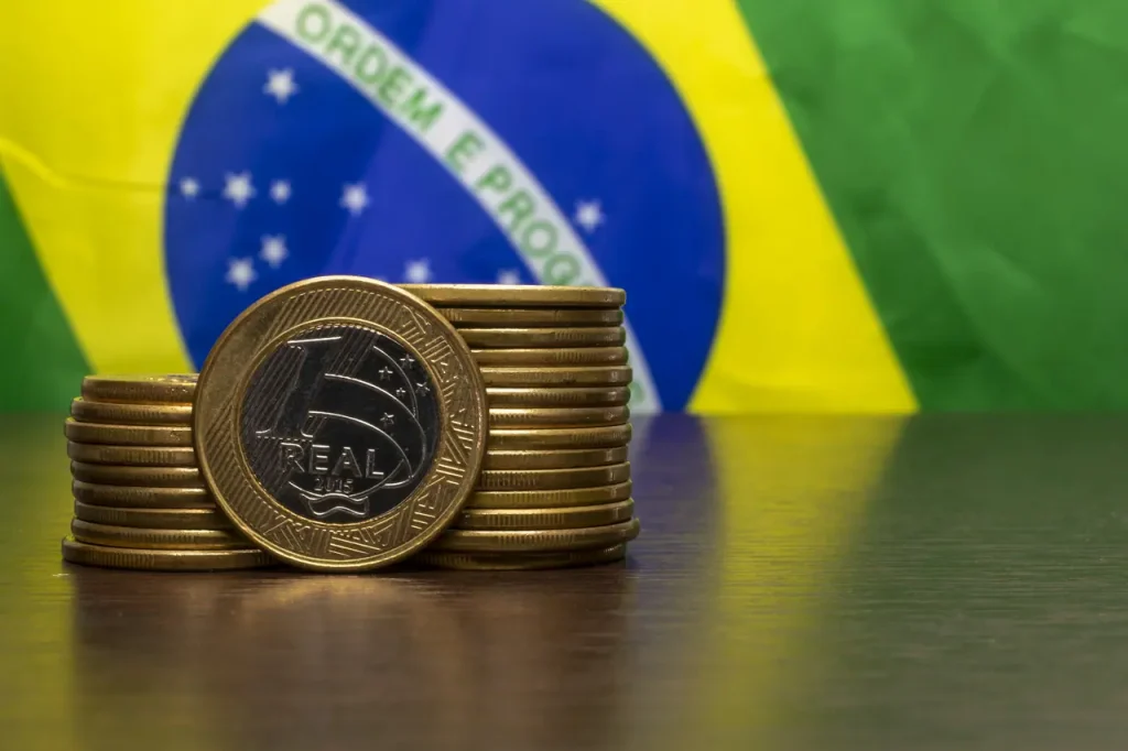 Imagem de moedas em frente à bandeira do Brasil, simbolizando a implementação do IBS e CBS na Reforma Tributária.