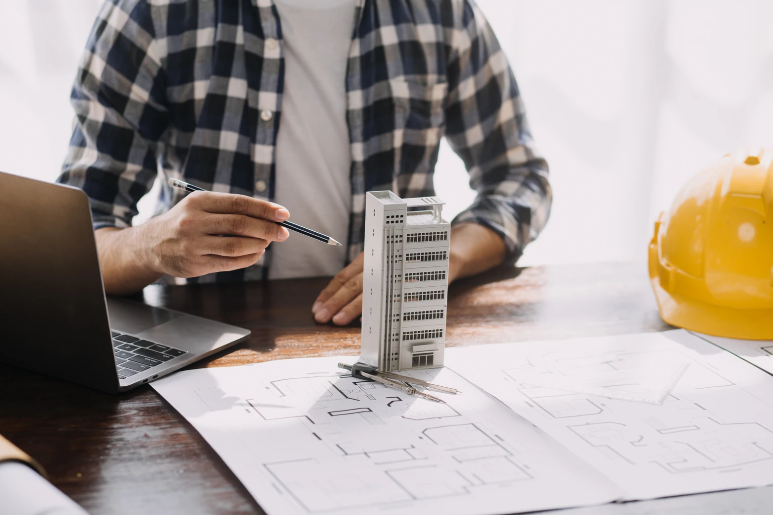 Profissional de construção civil analisando projeto de obra, ao lado de notebook e capacete de segurança